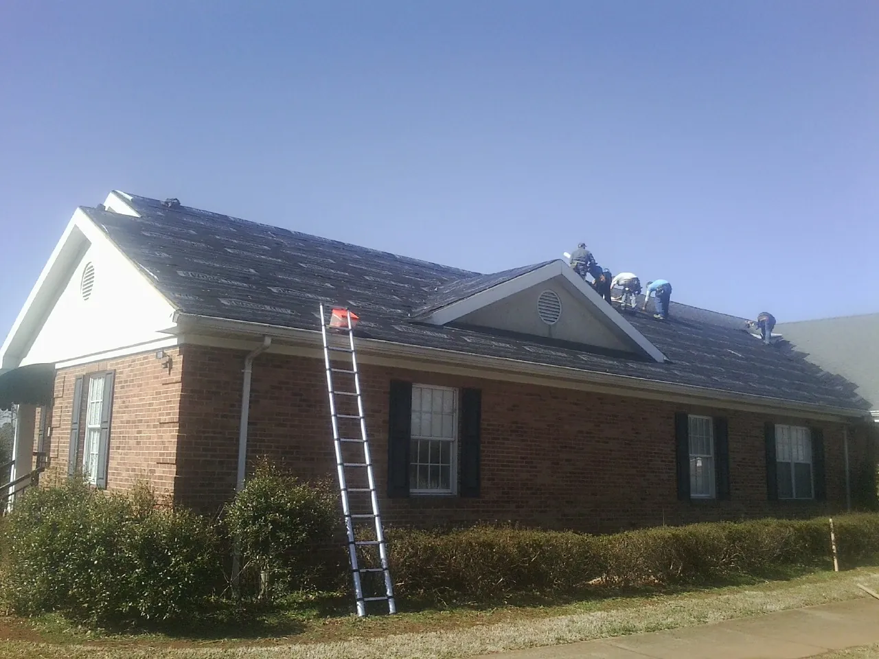 Crew tearing shingles off a brick ranch roof with a ladder set — Metal Roof Repair work by our crew in Raymore