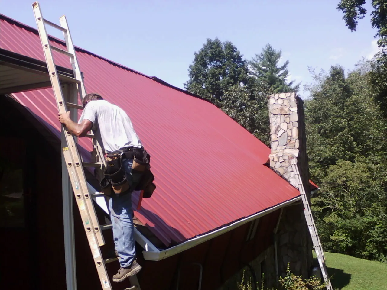 Roofer on a ladder beside a red metal roof and stone chimney — Soffit & Fascia Repair by Roofing Brothers in Raymore